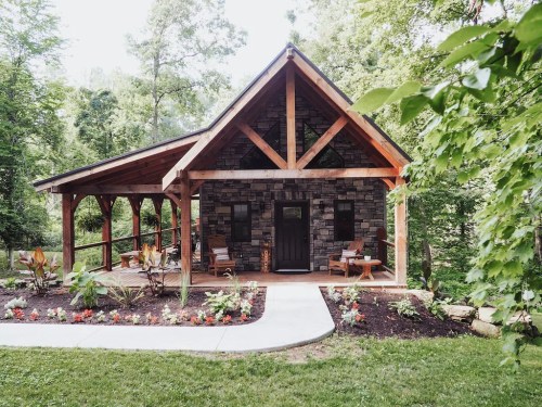 A charming stone cabin with a wooden porch, surrounded by greenery and colorful flowers.