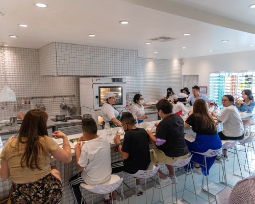 A busy restaurant interior with diners seated at a long counter, enjoying meals while chefs prepare food in the open kitchen.