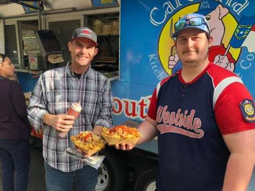 Two men hold plates of loaded nachos in front of a food truck, smiling and enjoying their meal.