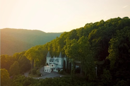 Aerial view of a large white mansion surrounded by lush green trees and mountains at sunset.