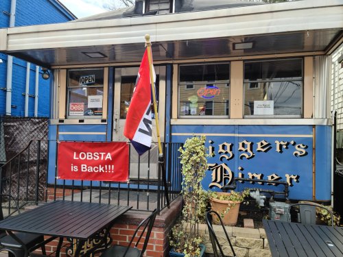 Exterior of a diner with a sign reading "LOBSTA is Back!!!" and a flag, featuring outdoor seating.