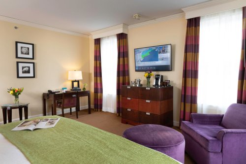 A cozy hotel room featuring a bed, purple armchair, TV, and a small table with flowers and coffee amenities.
