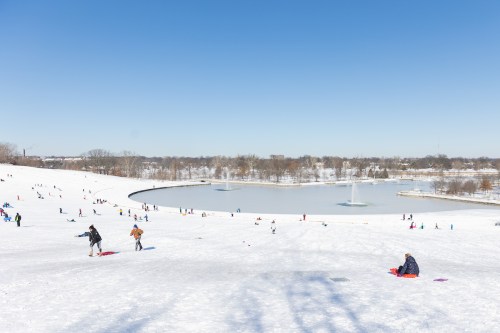 A snowy hill with people sledding, a frozen lake in the background, and clear blue skies above.