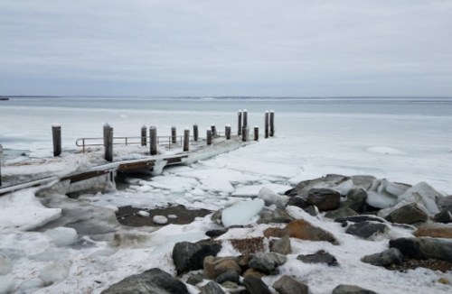 A frozen dock extends into a snowy, icy landscape with rocks in the foreground and a cloudy sky above.