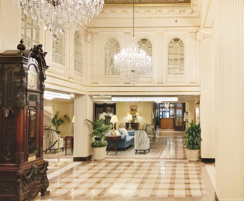 Elegant hotel lobby featuring chandeliers, ornate decor, and a vintage clock, with seating and plants.