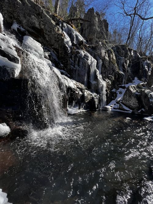 A serene waterfall cascades over rocky cliffs, surrounded by snow and ice under a clear blue sky.
