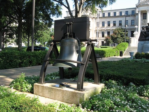 A large bell on a stone pedestal surrounded by greenery, with a building in the background.