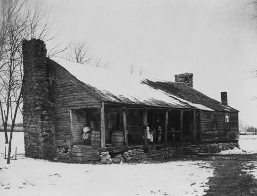 A historic wooden house with a snowy landscape, featuring a porch and two figures standing outside.