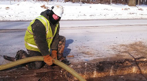 A worker in a safety vest and helmet uses a hose to remove water from a snowy construction site.