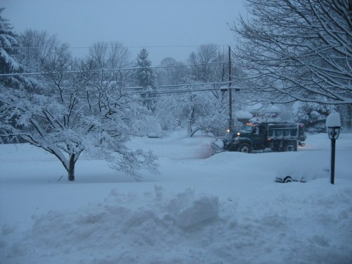 A snow-covered street with a snowplow clearing the road amidst trees blanketed in white snow.