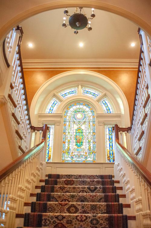 A grand staircase leads to a stained glass window, illuminated by soft lighting in an elegant interior.
