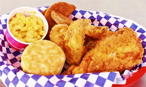 A basket of fried chicken, biscuit, macaroni and cheese, and potato wedges on a checkered paper liner.