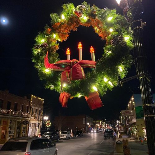 A festive wreath with lights and candles hangs on a street lamp, illuminating a charming nighttime scene.