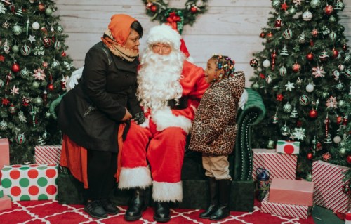 A woman and a girl in festive attire sit with Santa Claus in a decorated holiday setting with Christmas trees and gifts.