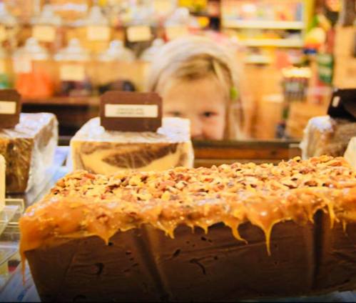 A child peeks from behind a display of colorful, assorted desserts in a shop.