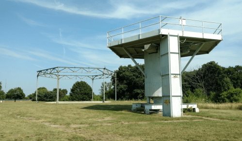 Two large, industrial structures on a grassy field, surrounded by trees under a clear blue sky.