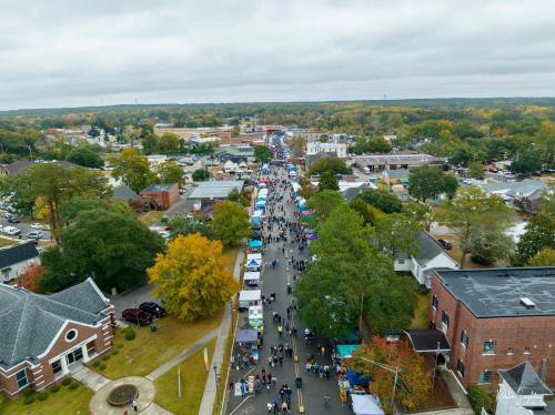 Aerial view of a bustling street fair with colorful tents, surrounded by trees and buildings under a cloudy sky.