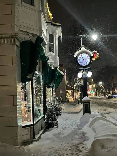 A snowy street scene at night, featuring a decorative clock and shop windows adorned with holiday decorations.
