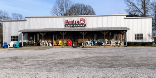 A rustic café-bakery with a wooden facade and colorful chairs outside, set in a gravel lot.