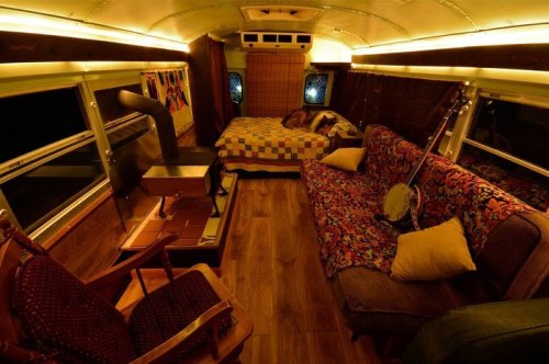 Cozy interior of a converted bus featuring a bed, sofa, rocking chair, and a banjo, illuminated by warm lighting.