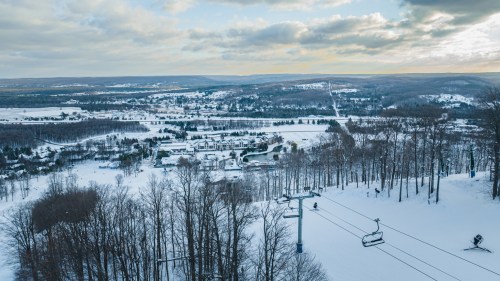A snowy landscape with a ski lift, trees, and distant hills under a cloudy sky.