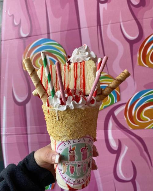 A colorful ice cream treat topped with whipped cream, candy, and cookies, held against a pink candy-themed background.