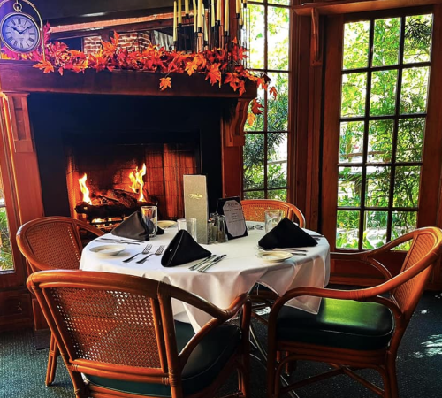 A cozy dining table set for two by a fireplace, surrounded by autumn decorations and large windows.
