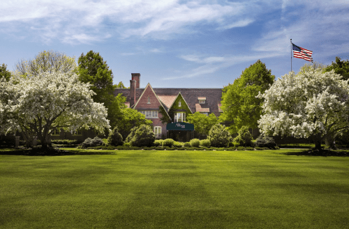 A large green lawn in front of a historic building, framed by flowering trees and an American flag waving in the background.