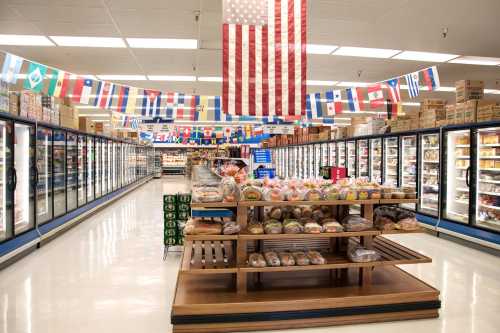 A grocery store aisle with freezers, a display of baked goods, and flags from various countries hanging overhead.