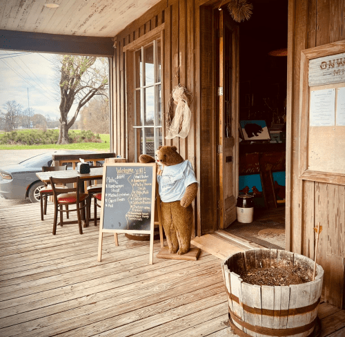 A bear statue in a blue shirt stands by a wooden entrance with a chalkboard sign and rustic decor.