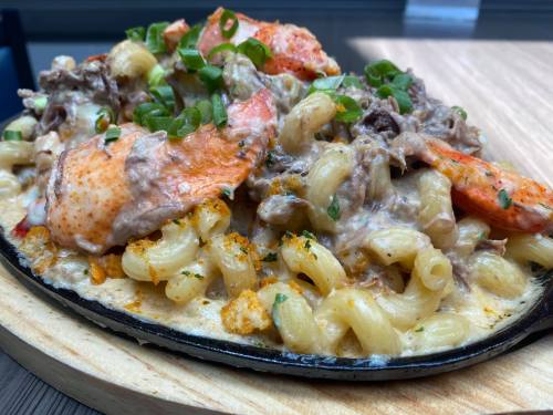 A sizzling platter of creamy pasta topped with lobster, beef, and green onions, served on a wooden board.