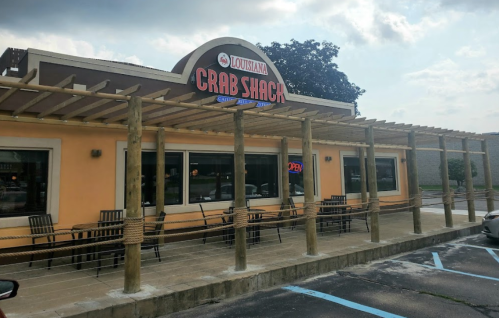 Exterior of Louisiana Crab Shack with a wooden pergola, outdoor seating, and an "Open" sign. Cloudy sky above.