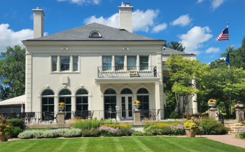 A large, elegant house with a balcony, surrounded by lush gardens and blue skies, featuring an American flag.