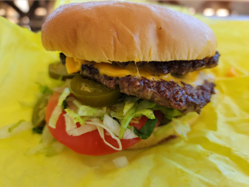 A close-up of a cheeseburger with lettuce, tomato, jalapeños, and a soft bun on yellow paper.
