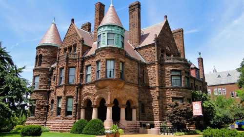 A large, historic stone mansion with turrets and a copper roof, surrounded by greenery and blue skies.