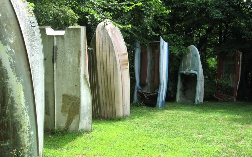A row of old, weathered boat hulls standing upright in a grassy area, surrounded by trees.