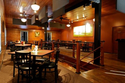 Cozy restaurant interior with wooden walls, tables, and chairs, featuring warm lighting and a staircase.