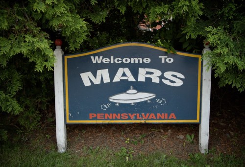A sign reading "Welcome to Mars, Pennsylvania" surrounded by greenery, featuring a UFO graphic.