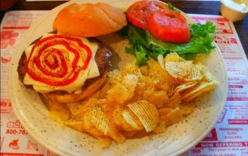 A plate with a burger topped with cheese and ketchup, served with lettuce, tomato, and a side of potato chips.