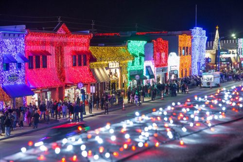 Colorful holiday lights adorn buildings along a bustling street, with crowds enjoying the festive atmosphere at night.