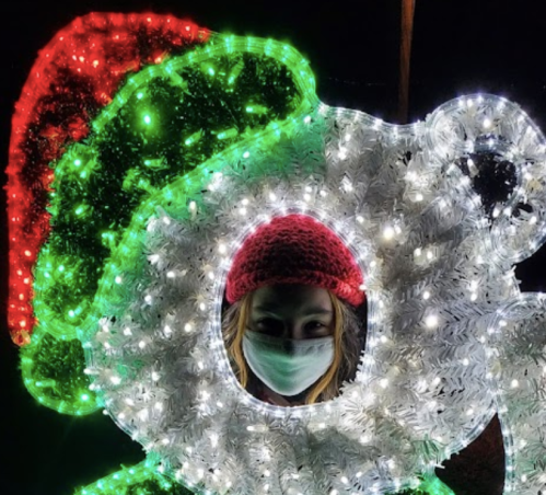 A person wearing a mask and a red hat peeks through a festive, illuminated decoration.