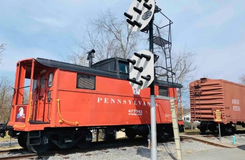 A red Pennsylvania caboose on display next to a railroad crossing sign and a freight car in a scenic outdoor setting.