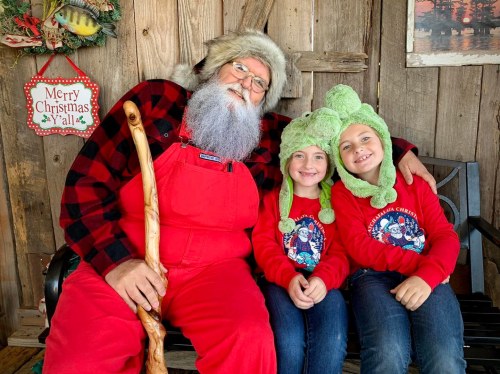 A bearded man in a red outfit sits with two girls wearing festive hats, smiling in a cozy holiday setting.