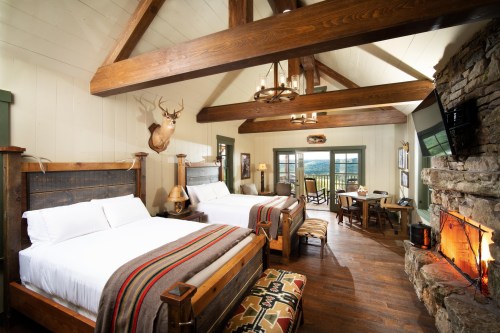 Cozy rustic bedroom with two beds, wooden beams, a stone fireplace, and a view of the outdoors.