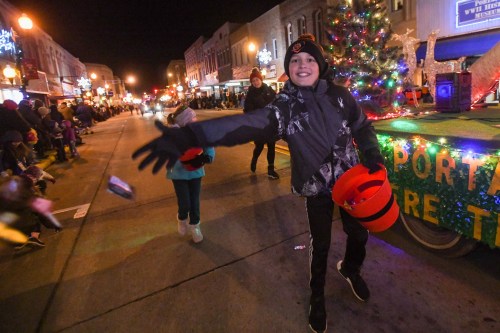 A child in winter attire throws candy from a float during a festive night parade, with holiday decorations in the background.