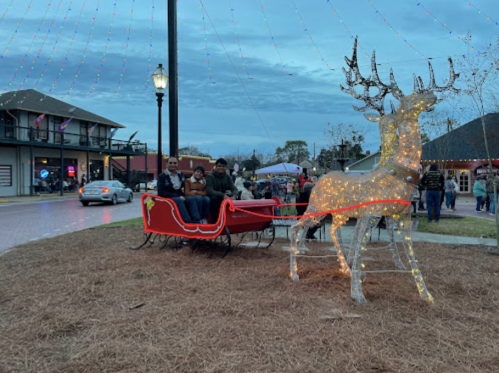 A festive scene with a lit reindeer and sleigh, surrounded by people and holiday decorations in a town square.