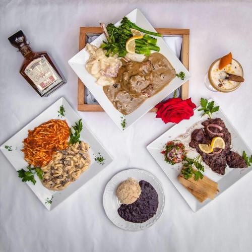 A beautifully arranged table with four gourmet dishes, a bottle of liquor, and a rose, set against a white background.