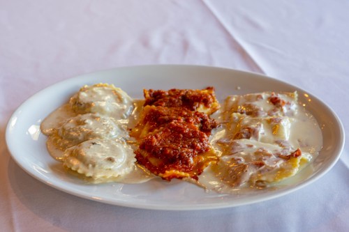 A white plate with ravioli and lasagna, topped with creamy sauce and meat sauce, on a white tablecloth.