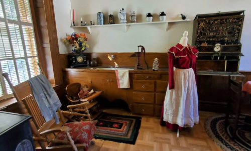 A vintage room featuring a rocking chair, a dress on a mannequin, and decorative items on shelves.