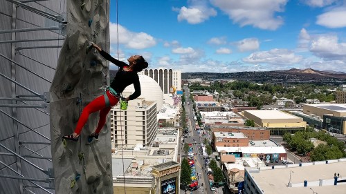 A climber ascends a tall rock wall with a cityscape and blue sky in the background.